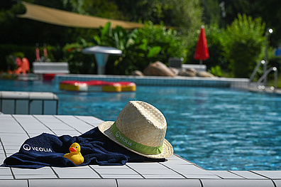 Poolside-Szenerie mit Strohhut mit grünem Band, dunkelbauem Handtuch mit VEOLIA-Logo und gelber Badeente auf grauer Poolkante. Im Hintergrund ein großes Schwimmbecken.
