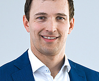 Ein Mann mit dunklen Haaren, der freundlich in die Kamera lächelt. Er trägt einen Anzug. 