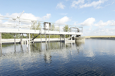 Ein großes, rundes Klärbecken einer Abwasseraufbereitungsanlage. Über das Becken spannt sich eine Räumerbrücke - eine lange, schmale Metallkonstruktion, die vom Rand bis zur Mitte des Beckens reicht.