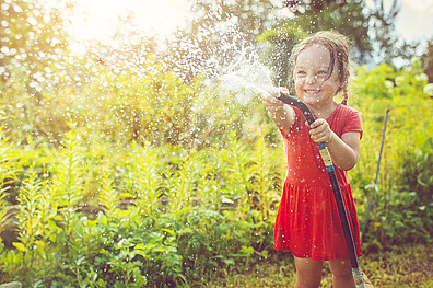 Ein Kind in einem roten Sommerkleid spielt an einem sonnigen Tag fröhlich mit einem Gartenschlauch. Umgeben von üppigem Grün sprüht das Wasser glitzernd durch die Luft, während die warme Sommersonne die Szene in ein goldenes Licht taucht.