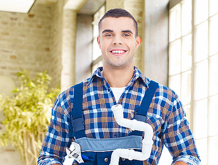 Ein junger Mann in Arbeitskleidung,kariertes Hemd und blaue Latzhose, steht für Installationsarbeiten bereit. In seiner rechten Hand hält er eine Wasserrohrszange, während er in der linken Hand ein Wasserrohr trägt.