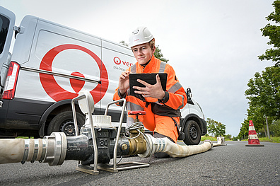 Ein Veolia-Mitarbeiter steht an einem Hydranten, einer Wasserentnahmestelle am Straßenrand. Er trägt Arbeitsschutzkleidung bestehend aus einem weißen Helm und einer orangefarbenen Jacke, die ihn gut sichtbar macht. Der Arbeiter führt gerade eine Durchflussmessung durch, um zu überprüfen, wie viel Wasser pro Zeiteinheit durch den Hydranten fließen kann.