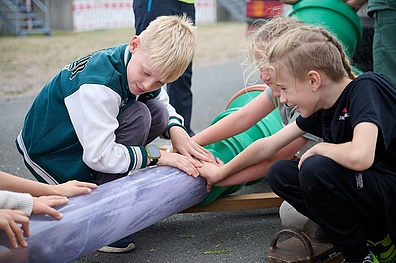 Drei Kinder im Grundschulalter bestauen eine Wasserrohrleitung. Sie haben ihre Hände drauf gelegt um die durchfließende Kraft des Wassers zu spüren.