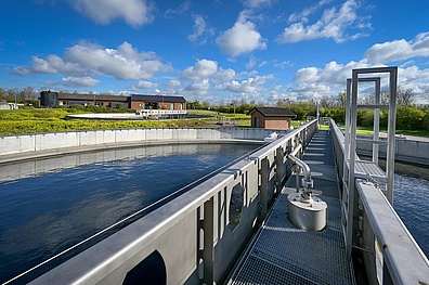 Blick von der Räumerbrücke auf das Aufbereitungsbecken der Kläranlage Schönebeck. Im Vordergrund ist der Metallsteg der Brücke zu sehen, im Hintergrund Betriebsgebäude und grüne Landschaft unter blauem Himmel mit weißen Wolken.
