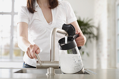 Eine Frau steht am Waschbecken. Sie macht Wasser in einen Wasserkocher.