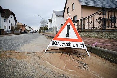 Wasserrohrbruch im Wohngebiet Am Straßenrand steht ein Warndreieck mit der Aufschrift: Achtung Wasserrohrbruch. Das Dreieck ist rot umrandet und hat in der Mitte ein schwarzes Ausrufezeichen auf weißem Grund.