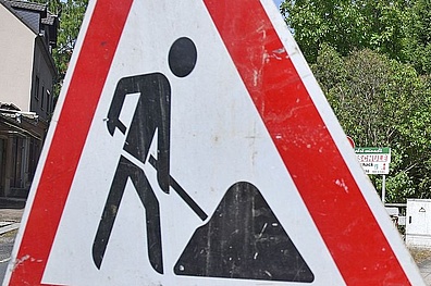 Ein dreieckiges Baustellenschild mit rotem rand zeigt das Symbol eines Bauarbeiters mit einer Schaufel.