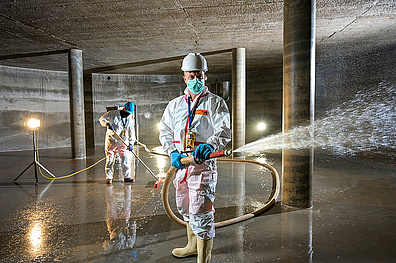 Zwei Mitarbeiter in weißen Schutzanzügen führen eine Reinigung in einem Hochbehälter für Trinkwasser durch. Sie stehen in dem großen, leeren Behälter und halten einen Wasserschlauch. Die Schutzkleidung und die Umgebung unterstreichen die Wichtigkeit der Hygiene bei der Trinkwasseraufbereitung.