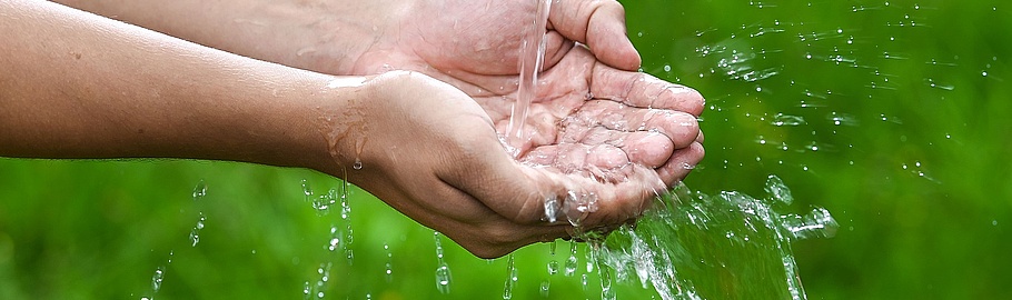 Zwei geöffnete Handflächen, nach oben gerichtet und leicht gewölbt, fangen klares Wasser auf. Die Hände bilden eine Schale, in der sich das Wasser sammelt.