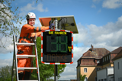 Ein Techniker in orangefarbener Schutzkleidung und weißem Schutzhelm steht auf einer Leiter und wartet eine digitale Geschwindigkeitsanzeige. Die Anzeigetafel zeigt 'SIE FAHREN' und einen digitalen Wert in km/h an. Im Hintergrund sind Wohnhäuser und Bäume zu sehen.