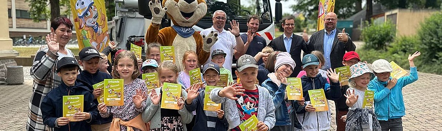 Eine Gruppe von Kindern steht auf einem gepflasterten Platz und hält gelbe Urkunden in die Kamera. Im Hintergrund ist das Maskottchen von 'Blicki blickt's' in Form eines großen Hundes sowie gelbe Werbefahnen der Initiative zu sehen. Die Kinder präsentieren stolz ihre Urkunden und winken in die Kamera, was auf eine Abschlussveranstaltung oder Ehrung im Rahmen des 'Blicki blickt's' Programms hindeutet.