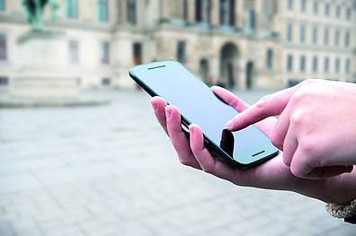 Nahaufnahme einer Hand die ein Smartphone in der Hand hält. Im Hintergrund ist verschwommen ein historisches Gebäude zu erkennen.
