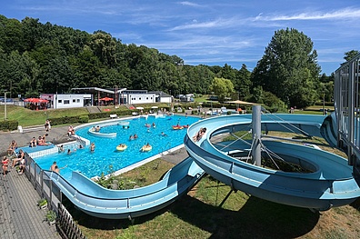 Luftaufnahme des Waldbads Colditz mit großem Schwimmbecken, Wasserrutschen, Besuchern auf Liegewiesen und umgebenden Grünanlagen an sonnigem Sommertag.