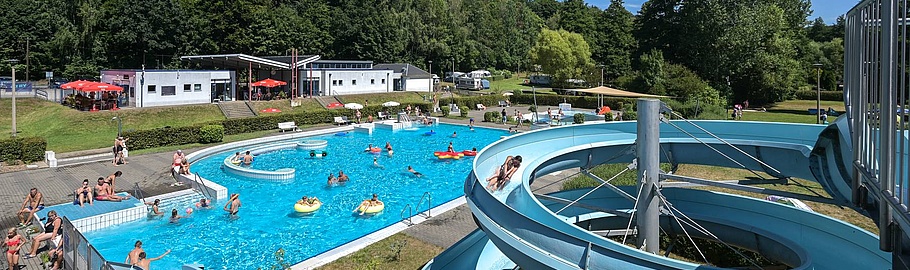 Luftaufnahme des Waldbads Colditz mit großem Schwimmbecken, Wasserrutschen, Besuchern auf Liegewiesen und umgebenden Grünanlagen an sonnigem Sommertag.