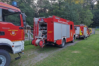 Drei rote Feuerwehrfahrzeuge hintereinander aufgereiht. Die Fahrzeuge stechen farblich deutlich gegen das umgebende Grün von Wald und Wiesen hervor.