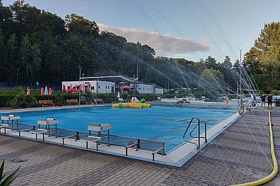 Das Schwimmbecken des Freibad Waldbad Colditz mit klarem blauen Wasser. Daneben liegt ein großer gelber Feuerwehrschlauch, aus dem viele vereinzelte Strahlen mit Wasser hinauslaufen.