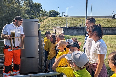 Auf dem Gelände der Kläranlage steht links ein Veolia-Mitarbeiter mit zwei Trichtern in den Händen – einer mit schmutzigem und einer mit sauberem Wasser. Er demonstriert die Abwasserreinigung. Gegenüber stehen Grundschulkinder mit ihrer Lehrkraft sowie einem weiteren Veolia-Mitarbeiter und hören aufmerksam zu.