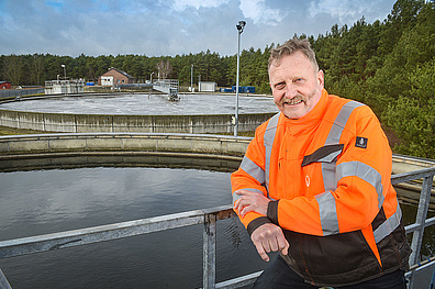 Ein Mitarbeiter in leuchtend orangefarbener Sicherheitskleidung steht lächelnd vor einer Kläranlage. Im Hintergrund sind mehrere runde Klärbecken zu sehen, umgeben von grünen Bäumen.