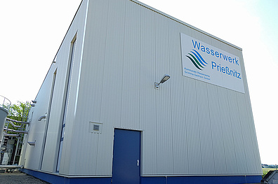 Ein großes, weißes Industriegebäude mit Wellblechfassade. An der Vorderseite ist ein blaues Logo mit der Aufschrift Wasserwerk Prisnitz zu sehen. Das Gebäude hat eine blaue Eingangstür und eine Außenbeleuchtung.
