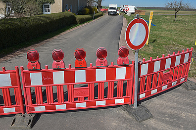 Eine Straße mit einer rot-weißen Baustellenabsperrung, die quer über die Straße verläuft. Davor steht ein Verkehrsschild, das ein Durchfahrtsverbot anzeigt.