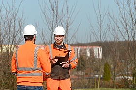 Zwei Mitarbeiter von Veolia in Arbeitskleidung und Schutzhelmen stehen sich gegenüber. Einer der Mitarbeiter hält ein Tablet in der Hand.