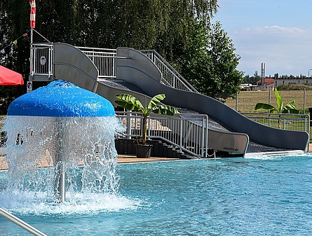 Ein Schwimmbecken mit einer grauen Wasserrutsche und einem blauen Wasserpilz, der Wasser sprüht. Am Beckenrand steht ein roter Sonnenschirm und einige Palmen in Kübeln sorgen für tropisches Flair.