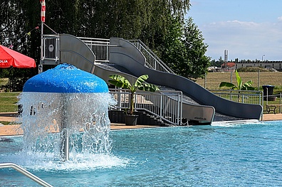 Ein Schwimmbecken mit einer grauen Wasserrutsche und einem blauen Wasserpilz, der Wasser sprüht. Am Beckenrand steht ein roter Sonnenschirm und einige Palmen in Kübeln sorgen für tropisches Flair.