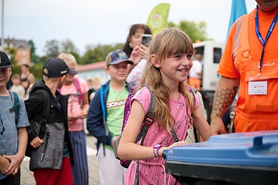 Ein Mädchen im Grundschulalter steht lächelnd mit einer großen Mülltonne, die sie mit den Händen festhält. Im Hintergrund sind weitere Kinder unscharf zu erkennen. Neben dem Mädchen befindet sich ein Veolia-Mitarbeiter in orangefarbener Arbeitskleidung.