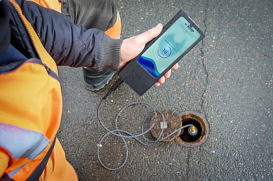 Nahaufnahme: Hand eines Veolia-Mitarbeiters hält den Alcom Leak-Tracker über einen geöffneten Messpunkt im Straßenboden.