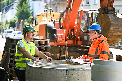 Zwei Mitarbeiter stehen auf einer Baustelle im Gespräch. Sie tragen Sicherheitskleidung, einer in einer gelben Warnweste mit grünem Helm, der andere in einer orangefarbenen Weste mit blauem Helm. Zwischen ihnen befindet sich ein großes Betonrohr.
