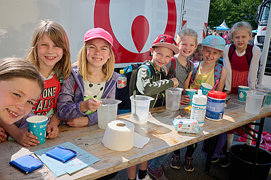 Das Bild zeigt eine Szene vom Internationalen Tag der Umwelt. Es sitzen mehrere Kinder an einem Tisch, auf dem verschiedene Utensilien für ein Umweltexperiment platziert sind.