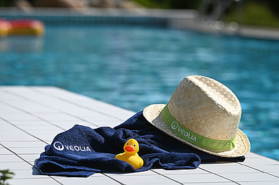 Am Rand eines Schwimmbeckens liegen ein dunkelblaues Handtuch mit Veolia-Logo, ein Strohhut mit grünem Veolia-Band und eine gelbe Badeente. Im Hintergrund ist das türkisfarbene Poolwasser mit schwimmenden roten Luftmatratzen zu sehen.