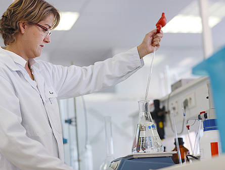 Eine Laborantin in einem weißen Kittel führt eine Trinkwasseranalyse durch. Sie steht in einem Labor und hantiert mit Präzisionsinstrumenten und Wasserproben.