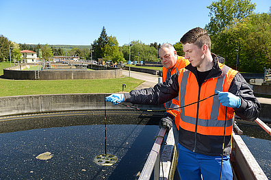Zwei Mitarbeiter in orangefarbenen Sicherheitswesten stehen an einem Geländer einer Kläranlage. Im Hintergrund sind Bäume und eine ländliche Umgebung zu sehen. Die Mitarbeiter von Veolia prüfen das Wasser.