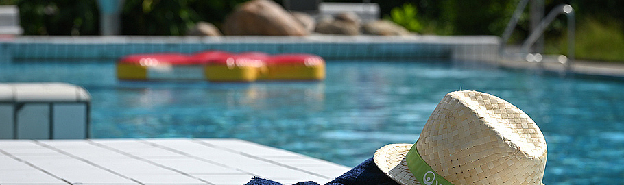 Im Vordergrund sitzt eine gelbe Badeente auf einem blauen Veolia Handtuch. Daneben liegt ein Strohhut mit einem grünen Veolia Logo-Band. Im Hintergrund ist ein mit klarem Wasser gefülltes Schwimmbecken in einem Freibad zu sehen.