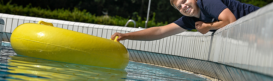 Ein junger Mann in einem dunkelblauen T-Shirt lehnt sich lächelnd über den Rand eines Schwimmbeckens und greift nach einer gelben aufblasbaren Luftmatratze, die auf dem Wasser treibt. Im Hintergrund sind grüne Bäume und Sträucher zu sehen. Die Szene zeigt eine entspannte Sommeratmosphäre im Freibad.