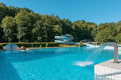 Das Bild zeigt das Waldbad Colditz. Im Zentrum des Bildes befindet sich ein großes Schwimmbecken mit strahlend blauem Wasser. Das Becken ist umgeben von einer üppigen, grünen Waldlandschaft. Zahlreiche Bäume säumen den Rand des Bades und verleihen ihm eine erholsame Atmosphäre.