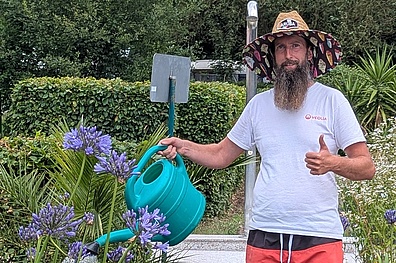 Mann mit Bart und gemustertem Hut gießt Pflanzen im Waldbad Colditz mit türkisfarbener Gießkanne. Lila Blüten und grüne Hecken im Hintergrund.
