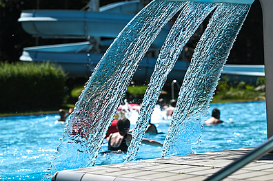 Drei kräftige Wasserstrahlen spritzen aus Düsen am Poolrand in ein blaues Schwimmbecken. Im Hintergrund sind verschwommen mehrere Badegäste im Wasser zu sehen.