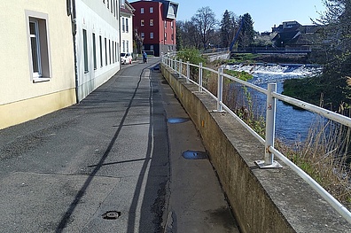 Blick entlang der Staupitzstraße in Döbeln: Ein Gehweg verläuft neben einem Fluss mit weißem Geländer