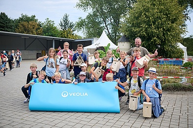 Ein Gruppenfoto zeigt eine Grundschulklasse mit zwei Lehrern an den Außenseiten der Gruppe. Viele der Kinder und die Erwachsenen strecken den Daumen nach oben in Richtung Kamera. Vier Jungen halten gemeinsam ein blaues Veolia-Banner vor sich.