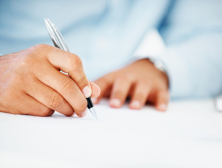 Nahaufnahme von zwei Händen. Die rechte Hand hält einen Stift und unterschreibt Dokumente.