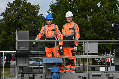 Tim Winkler (links) und Franz Neudahm (rechts) stehen auf einer erhöhten Metallplattform der Kläranlage Bad Lausick. Beide tragen orangefarbene Warnschutzkleidung mit reflektierenden Streifen sowie Schutzhelme - Winkler einen blauen und Neudahm einen weißen. Sie inspizieren die aus Metallkonstruktionen und blauen Komponenten bestehende technische Anlage. Im Hintergrund sind grüne Bäume zu sehen.