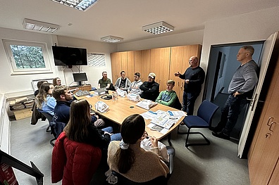 Ein Besprechungsraum mit einem großen Tisch, um den herum etwa 15 Personen sitzen oder stehen. Die Gruppe besteht hauptsächlich aus jungen Erwachsenen, mit einigen älteren Teilnehmern. An der Stirnseite des Tisches steht ein Mann, der zu der Gruppe spricht.