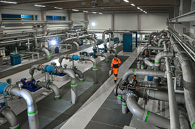 Innenansicht einer Wasseraufbereitungsanlage. Der Raum ist gefüllt mit einem Netzwerk aus silbernen Rohren, Pumpen und Tanks verschiedener Größen. Ein Mitarbeiter in orangefarbener Arbeitskleidung ist in der Mitte des Bildes zu erkennen.