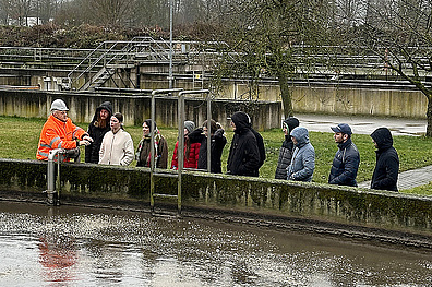Eine Gruppe von jungen Menschen steht am Rand eines Belebungsbeckens in einer Kläranlage. Ein Arbeiter in orangefarbener Sicherheitskleidung und Schutzhelm erklärt etwas, während die anderen aufmerksam zuhören. Die Zuhörer tragen warme Winterkleidung. Im Hintergrund sind kahle Bäume und industrielle Strukturen der Kläranlage zu sehen.