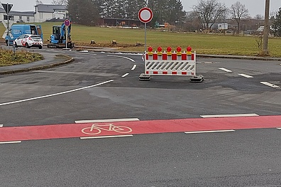 Leere, gesperrte Kreuzung mit rot-weißer Absperrung im Vordergrund, dahinter zwei parkende Autos und ein Bagger.