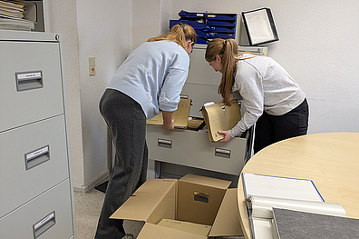 Zwei Mitarbeiterinnen in einem Büro packen Akten aus einem Aktenschrank in einen Umzugskarton.