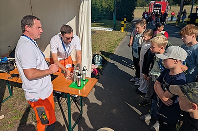 Veolia-Mitarbeiter aus dem Energiebereich stehen neben einem Tisch, auf dem ein Modell zur Veranschaulichung erneuerbarer Energien aufgebaut ist. Sie erklären den davor stehenden Grundschulkindern die Funktionsweise, während die Kinder aufmerksam zuhören.