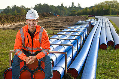 Martin Moisel, operativer Leiter bei Veolia, sitzt auf mehreren blauen Trinkwasserleitungen. Er trägt einen Schutzhelm und Arbeitskleidung.